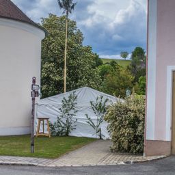Ein weißes Zelt steht in einem Hof mit einem Holzhocker daneben. Ein Gebäude mit einem braunen Dach befindet sich links, und ein Busch mit Blumen rechts. Bäume und ein bewölkter Himmel sind im Hintergrund.