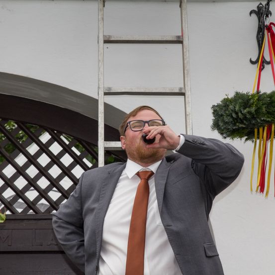 Ein Mann in einem grauen Anzug trinkt aus einem Glas und steht vor einer weißen Wand mit einem dekorativen Kranz und einer Leiter.