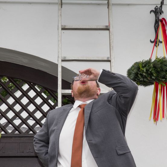 Ein Mann trinkt aus einem Glas, während er draußen steht und sich gegen eine weiße Wand mit einer Leiter und einem dekorativen Kranz lehnt.