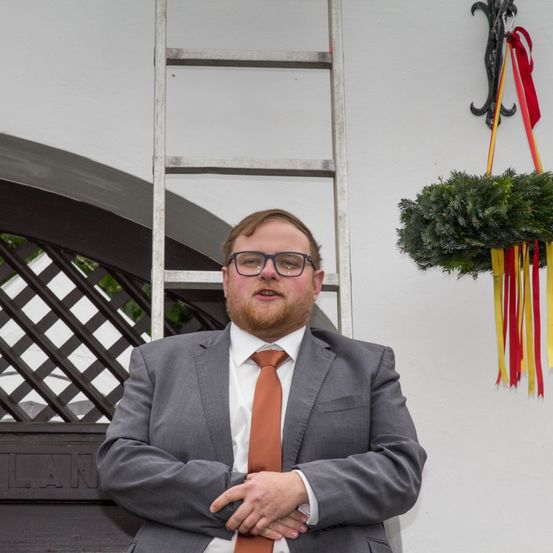 Ein Mann in Anzug und Krawatte steht mit verschränkten Armen vor einem Gebäude. Eine Leiter und ein Kranz hängen an der Wand.