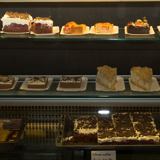 Eine Vitrine mit verschiedenen Desserts wie Kuchen und Torten, einige mit Früchten oder Schokolade garniert. Unten steht ein Schild 'Schwarzwalder-Kirsch-Torte'.