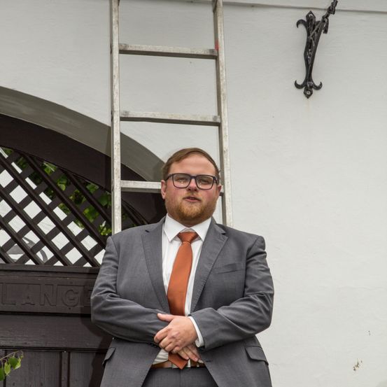 Ein Mann in einem grauen Anzug mit einer orangefarbenen Krawatte steht vor einem weißen Gebäude. Eine Leiter und ein schwarzes Metallobjekt sind ebenfalls zu sehen.