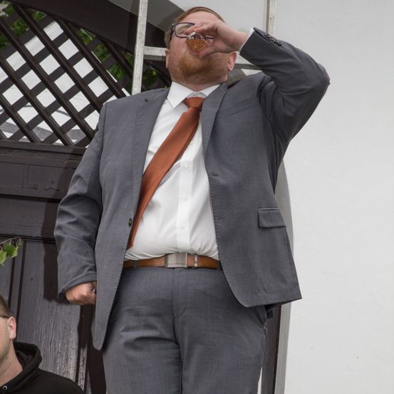 Ein Mann in einem grauen Anzug und einer orangefarbenen Krawatte trinkt aus einem Glas und steht auf einer Holztreppe.