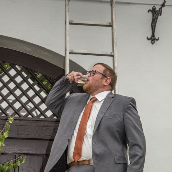 Ein Mann in einem grauen Anzug und einer orangefarbenen Krawatte trinkt aus einem Glas vor einem Gebäude mit einer Tür, die mit 'LANG' beschriftet ist, und einer Leiter an der Wand.
