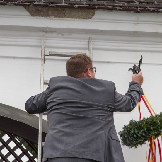 Ein Mann in einem grauen Anzug und Brille klettert eine Leiter hoch, um einen Kranz an einem Gebäude aufzuhängen.