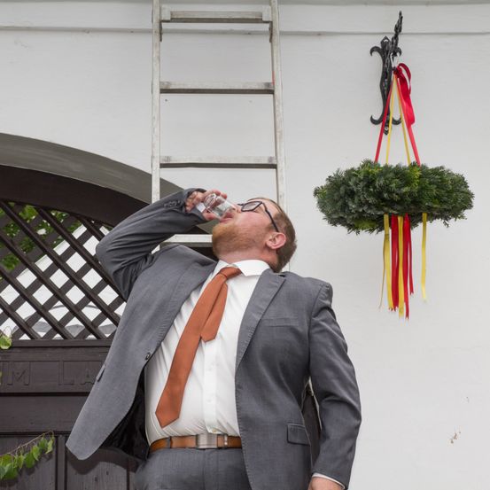 Ein Mann in einem grauen Anzug trinkt Wasser aus einer Flasche, während er nach oben schaut. Ein dekorativer Kranz hängt an der Wand hinter ihm.