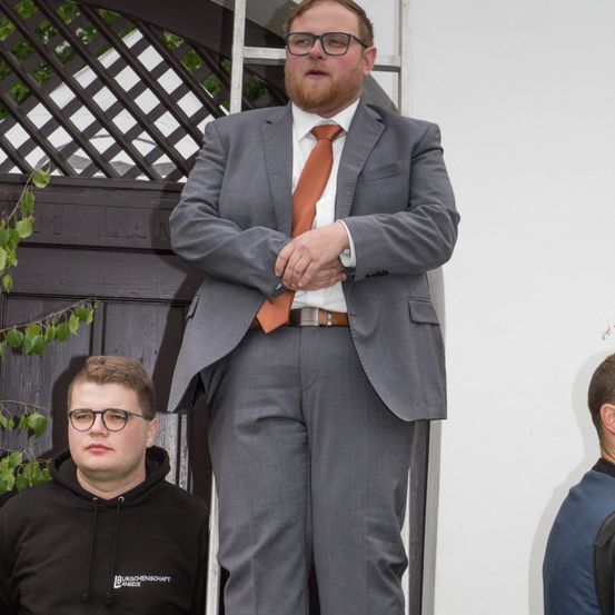 Ein Mann in einem grauen Anzug und einer orangefarbenen Krawatte steht auf einer Veranda und scheint ein Publikum anzusprechen. Er steht zwischen zwei anderen Männern, einem in einer schwarzen Kapuzenjacke und dem anderen in einem blauen Hemd. Hinter ihm befindet sich eine Holztür mit einem Spalier.