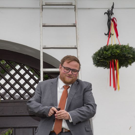 Ein Mann in einem grauen Anzug mit einer roten Krawatte hält ein Glas Wein vor einem Haus. Hinter ihm befinden sich eine Tür, ein Fenster, eine Leiter und ein Kranz.
