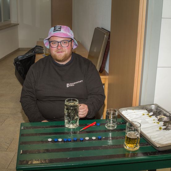 Ein Mann sitzt an einem Tisch, lächelt, mit einem Bierkrug und einem Brettspiel-Setup. Er trägt eine Batikmütze und eine Brille.