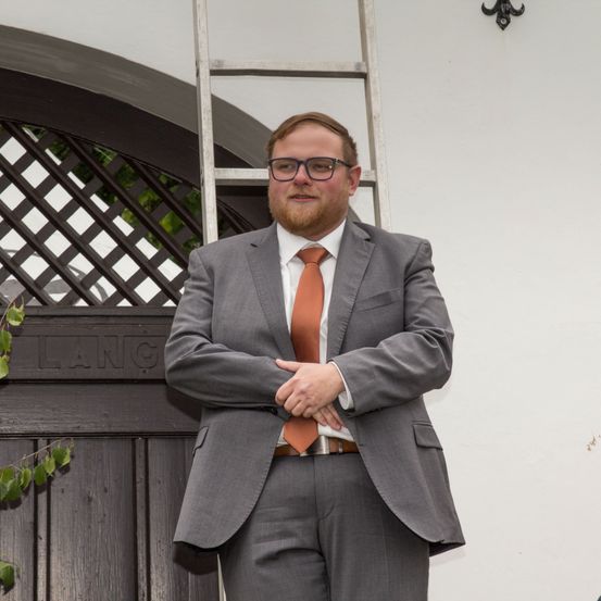 Ein Mann mit Bart, der einen grauen Anzug, eine orange Krawatte und eine Brille trägt, steht mit verschränkten Armen vor einer Holztür. Dahinter befindet sich eine weiße Wand mit einer Leiter und einem dekorativen Metallstück.