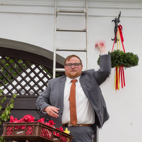 Ein Mann in einem grauen Anzug und einer orangefarbenen Krawatte steht vor einem Haus, lächelt und scheint eine Girlande aufzuhängen. Eine Leiter lehnt an der Wand, und es gibt zwei rote Blumenarrangements am Boden.