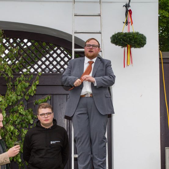 Ein Mann in formeller Kleidung steht auf einer Leiter, während ein anderer Mann in einer schwarzen Kapuzenjacke in der Nähe steht. Ein dritter Mann hält ein Glas in der Hand. Die Szene spielt sich vor einem Gebäude ab, an dem ein dekorativer Kranz an der Wand hängt.