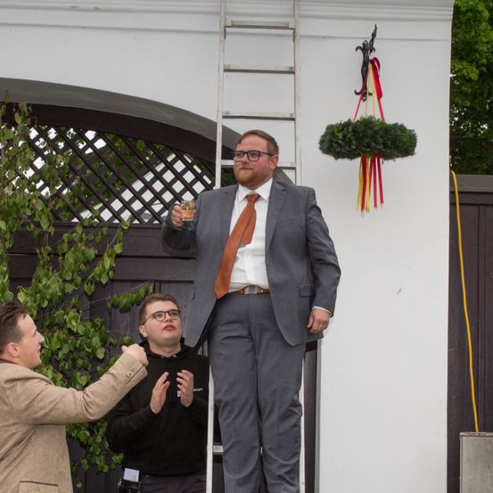 Ein Mann steht auf einer Leiter und hält ein Glas mit Flüssigkeit in der Hand, angezogen in einem grauen Anzug, während ein anderer Mann in der Nähe gestikuliert. Dahinter ist ein Baum mit grünen Blättern zu sehen.