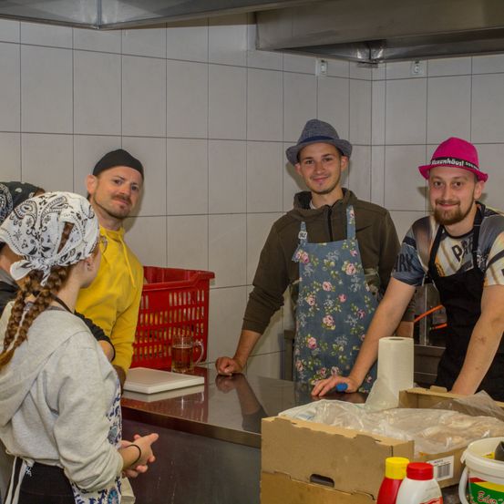 Fünf Personen befinden sich in einer Küche, jede trägt eine Schürze. Eine Frau mit Kopftuch steht links, ein Mann mit Hut neben ihr. Ein weiterer Mann in einem gelben Shirt steht links, während zwei Männer mit Hüten rechts sind.