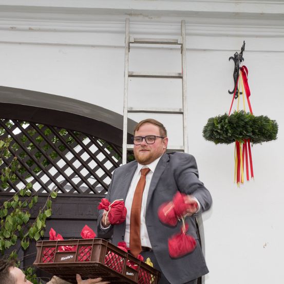 Ein Mann in Anzug hält einen Korb mit roten Geschenken auf einer Veranda. Eine Leiter und ein Kranz sind hinter ihm.