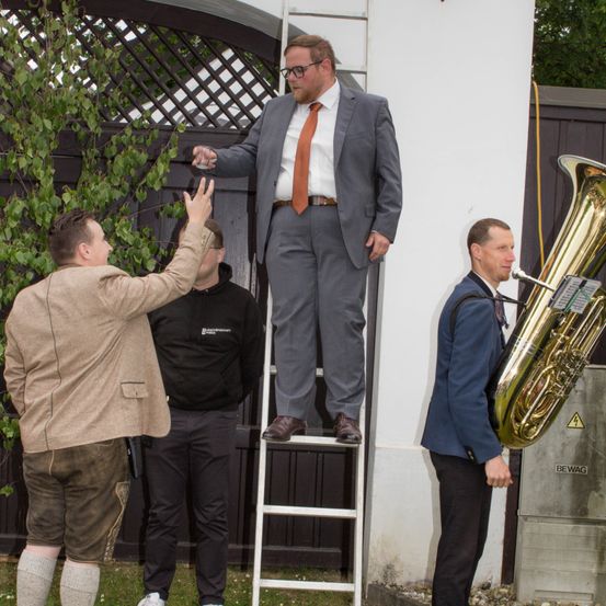 Ein Mann auf einer Leiter hebt ein Glas, während ein anderer Mann jubelt. Ein Musiker spielt Tuba in der Nähe. Sie sind draußen, möglicherweise bei einer Feier.