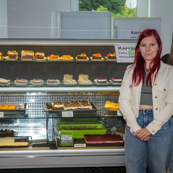 Eine Frau mit roten Haaren steht lächelnd vor einer Vitrine mit Desserts. Die Vitrine enthält verschiedene Kuchen und Gebäck. Über der Vitrine befindet sich ein Schild mit der Aufschrift 'Kaffee Mehlspeise'.