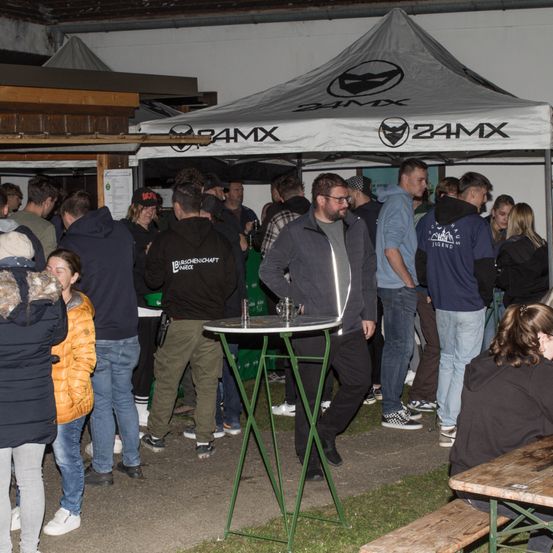 Eine Gruppe von Menschen steht um einen kleinen Tisch unter einem Zelt mit dem Logo 24MX. Die Leute sind in Jacken, Jeans und Turnschuhen gekleidet. Einige schauen sich um, während andere sich auf den Tisch konzentrieren.