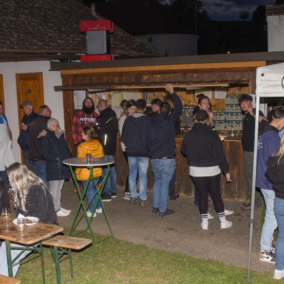 Menschen sind um einen Imbissstand versammelt, einige stehen, andere sitzen auf Bänken. Ein Mann in einem schwarzen Hoodie steht vor der Theke und hält ein Glas. Einige der Leute schauen sich das Menü an, während andere in Gespräche vertieft sind.