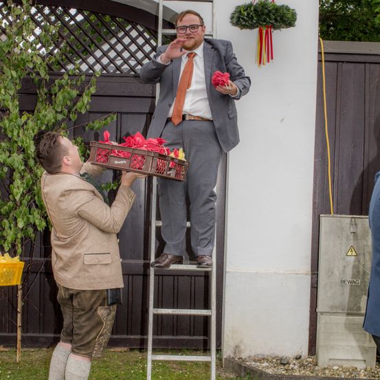 Ein Mann in formeller Kleidung steht auf einer Leiter und hält einen Blumenstrauß, während ein anderer Mann unten einen Korb mit Blumen hält. Sie sind von Grünpflanzen und einem Holzzaun umgeben.