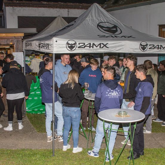 Eine Gruppe von Menschen versammelt sich unter einem 24MX-Zelt, einige stehen und andere sitzen auf Stühlen um einen Tisch, genießen Getränke. Der Bereich ist im Freien mit Zelten und einem Gebäude im Hintergrund.