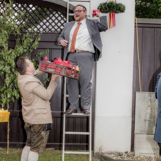 Ein Mann in Anzug klettert eine Leiter hoch und hält ein rotes Band, während ein anderer Mann in traditioneller Kleidung einen Korb mit roten Bändern hält. Ein Zaun und eine Pflanze mit grünen Blättern sind im Hintergrund.