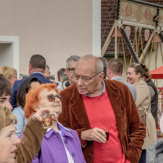 Ein Treffen älterer Menschen im Freien; ein Mann in einem roten Hemd und eine Frau mit roten Haaren halten Getränke. Mehrere Menschen unterhalten sich, und ein Gebäude mit aufwendigen Verzierungen ist im Hintergrund.