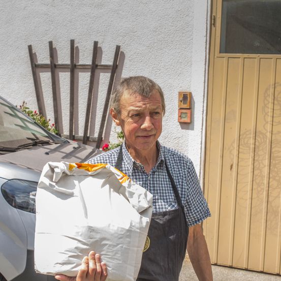 Ein älterer Mann in einem karierten Hemd und einer Schürze hält einen weißen Beutel vor einem Haus mit einem Auto und einem Holzzaun.