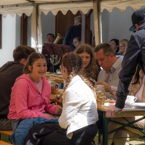 Eine Gruppe von Menschen sitzt draußen an einem Tisch, vielleicht auf einem Festival. Zwei Frauen lächeln, während sie nebeneinander sitzen. Ein Mann vor ihnen sieht nach unten. Ein Mann in einer schwarzen Jacke steht neben ihnen.