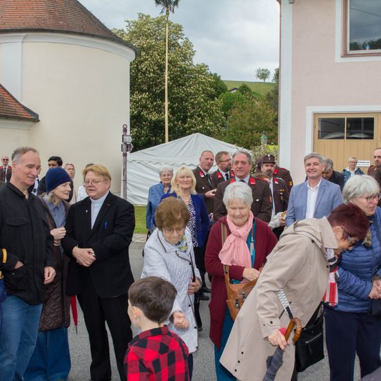 Eine Gruppe von Menschen, einschließlich älterer Personen und Kindern, versammelt sich vor einem Gebäude. Einige tragen formelle Kleidung, während andere in Freizeitkleidung sind. Die Szene umfasst ein Gebäude mit einem braunen Dach, einem Fahnenmast und einem weißen Zelt.