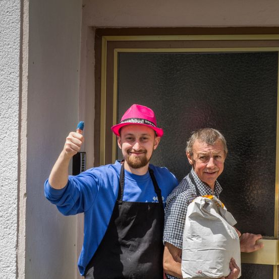 Ein Mann mit rosa Hut gibt einen Daumen hoch und ein anderer Mann hält einen Beutel mit Essen vor einem Gebäude.