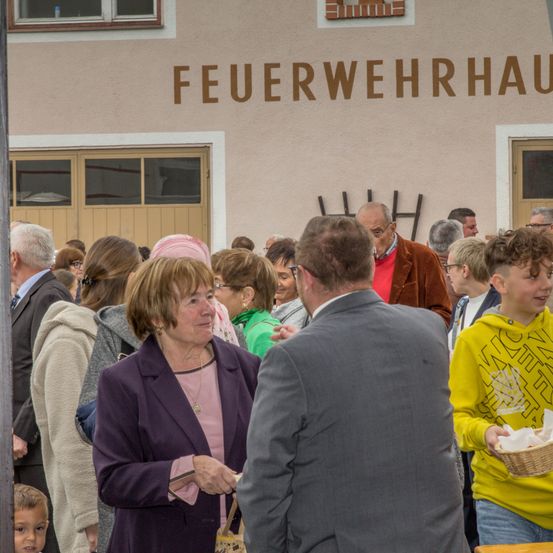 Eine Menschenmenge, darunter eine ältere Frau, steht vor einem Gebäude mit der Aufschrift 'Feuerwehrhaus'. Sie scheinen zu unterhalten.