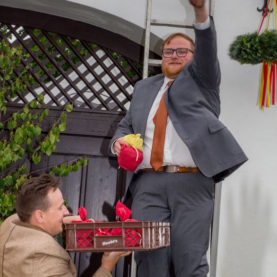 Ein Mann mit Brille und Anzug steht auf einer Leiter und hält ein rot-gelb verpacktes Objekt, während ein anderer Mann, der einen Korb mit verpackten Gegenständen hält, ihn anlächelt.