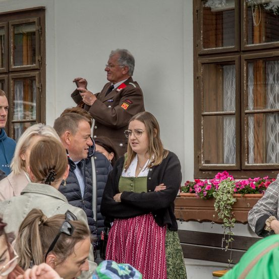 Ein Mann in Uniform macht ein Foto mit einer Kamera. Vor ihm steht eine Frau mit grünem Schürzen und verschränkten Armen. Dahinter unterhalten sich mehrere Menschen. Im Hintergrund ein Fenster mit rosa Blumen.