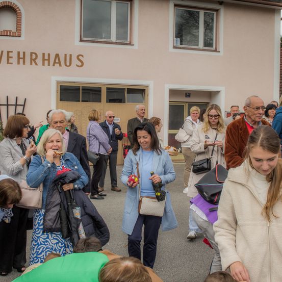 Eine Gruppe von Menschen steht vor einem Gebäude mit der Aufschrift 'Verrhaus'. Einige essen, während andere Taschen tragen. Eine Frau hält Blumen und eine Flasche. Ein junges Mädchen schaut nach unten.