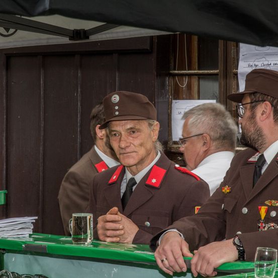 Zwei ältere Männer in Uniform sitzen an einem grünen Tresen, einer mit einem Glas, andere dahinter. Im Hintergrund ist eine Holztür und Papiere zu sehen.