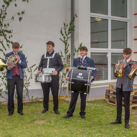 Fünf junge Männer in Uniformen spielen Blechblas- und Schlagzeuginstrumente auf einer Rasenfläche vor einem Gebäude.