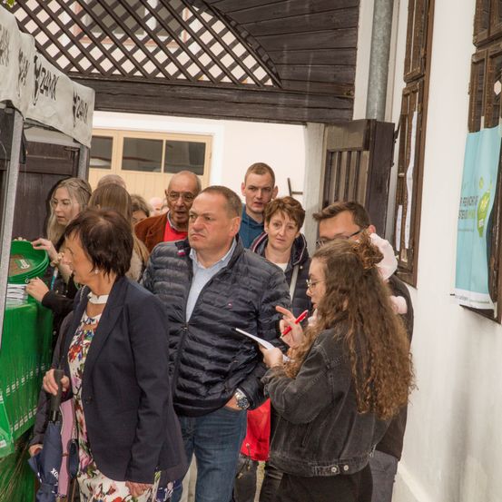 Eine Gruppe von Menschen versammelt sich in einem Korridor, einige schreiben auf Papiere, andere unterhalten sich. Im Hintergrund sind grüne Lagerboxen und ein Plakat zu sehen.