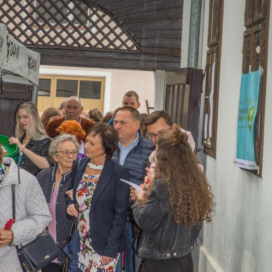 Mehrere Menschen sind in einem Außenbereich versammelt, wahrscheinlich bei einer Veranstaltung. Sie stehen in einer Schlange und unterhalten sich miteinander. Einige von ihnen tragen Brillen und haben Handtaschen dabei. Ein Plakat ist an der Wand befestigt.