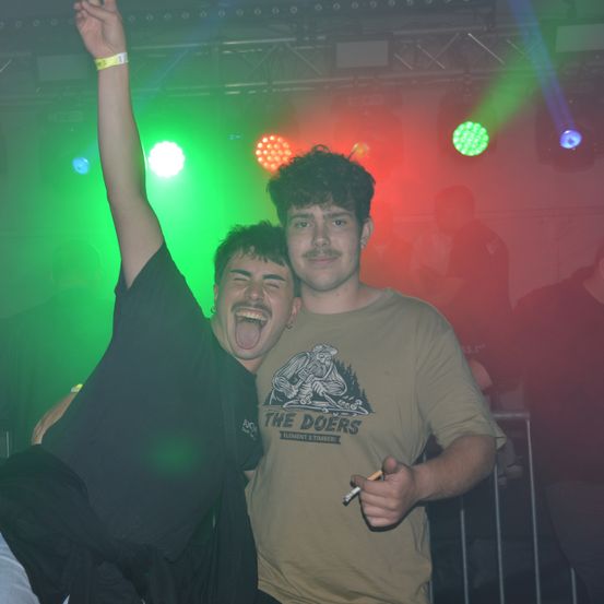 Zwei junge Männer stehen in einem Club, einer mit erhobenem Arm und der andere raucht eine Zigarette. Sie stehen unter grünem und rotem Licht.