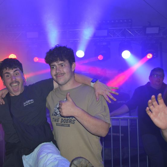 Zwei junge Männer lächeln und posieren für ein Foto in einem Nachtclub. Einer gibt einen Daumen hoch und beide sind von farbigen Lichtern umgeben.