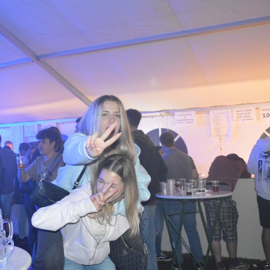 Zwei junge Frauen posieren für ein Foto in einem Club, eine trägt eine weiße Kapuzenjacke und die andere eine blaue Kapuzenjacke. Sie machen Friedenszeichen. Ein Tisch mit Gläsern und eine Person, die sich darauf lehnt, ist im Hintergrund.