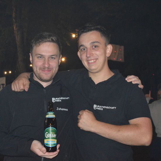 Zwei Männer in schwarzen Jacken posieren bei Nacht für ein Foto, einer hält eine Flasche Gösser Bier, mit verwaschenem Hintergrund und festlicher Atmosphäre.