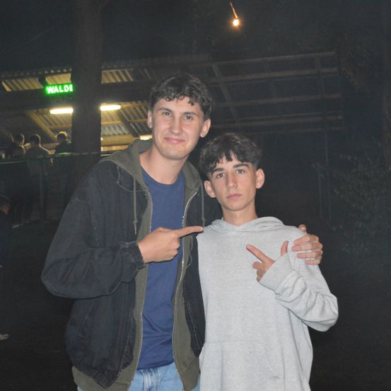 Zwei junge Männer stehen zusammen in einem schwach beleuchteten Bereich, einer zeigt mit seiner rechten Hand. Sie lächeln beide, tragen Jacken und stehen unter einem Schild, das 'WALDE' steht.