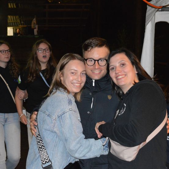 Fünf Menschen stehen nachts in einer Gruppe. Zwei Frauen und ein Mann lächeln und posieren für ein Foto. Dahinter singt eine Frau.