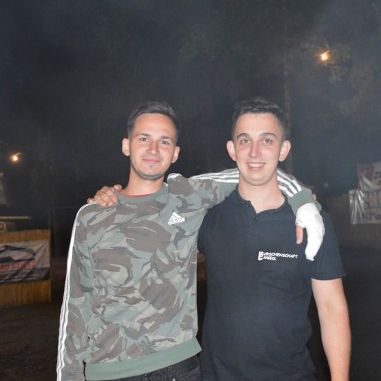 Zwei Männer stehen nachts draußen. Einer trägt ein Camouflage-Shirt, der andere ein schwarzes Polohemd mit weißem Text. Sie lächeln für die Kamera.