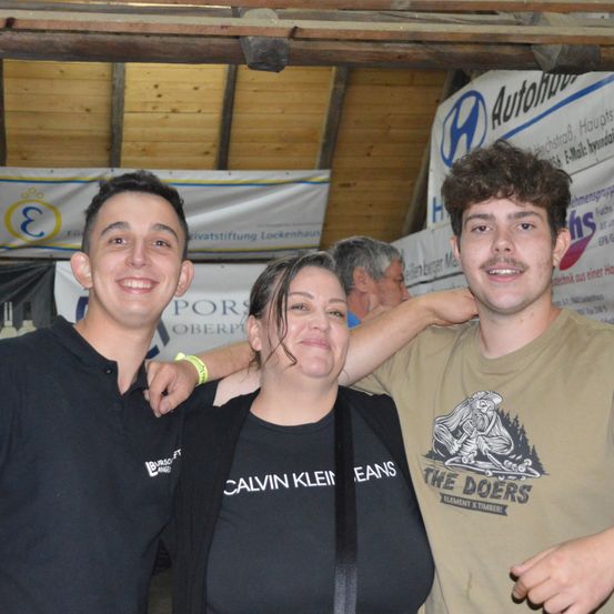 Drei Personen lächeln für ein Foto. Eine Frau und zwei Männer, einer in einem Calvin Klein-Shirt und der andere in einem The Doers-Shirt. Dahinter ist ein Banner mit 'Autohaus' und 'PORSCHE OBERPOLLI'.