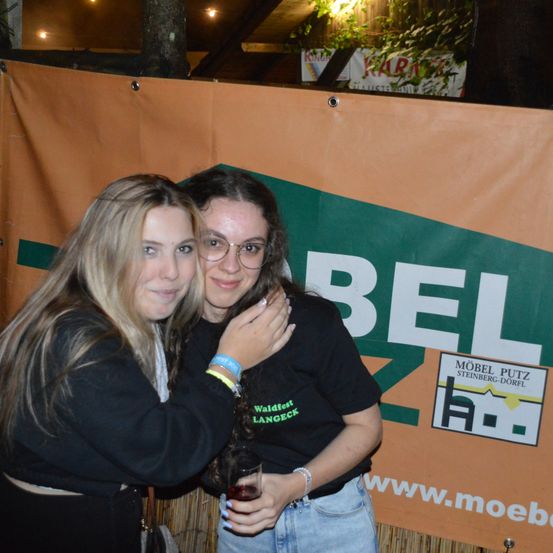 Zwei Frauen posieren zusammen vor einem Banner. Das Banner hat grüne, weiße und orangefarbene Farben mit dem Text 'BEL'. Die Frau auf der linken Seite hat lange blonde Haare und trägt eine Brille. Die Frau auf der rechten Seite trägt ein Armband und hält ein Glas.