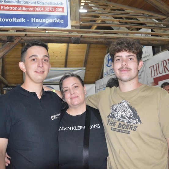 Drei Menschen posieren für ein Foto in einem Innenraum, wahrscheinlich bei einer Veranstaltung. Zwei Männer stehen auf beiden Seiten einer Frau und lächeln für die Kamera. Hinter ihnen ist eine Holzkonstruktion an der Decke befestigt. Ein weißes Banner mit Text hängt an der Decke.