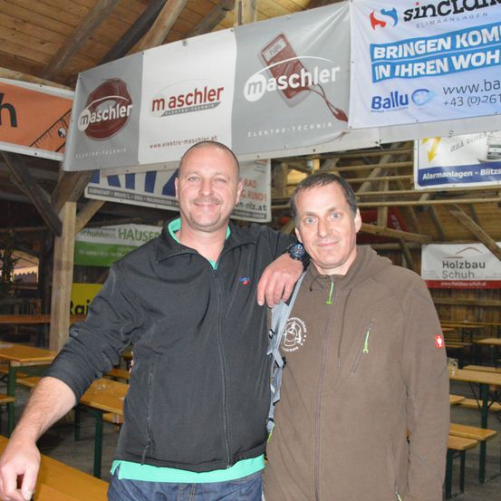 Zwei Männer stehen in einem Holzgebäude mit einem Banner hinter ihnen. Sie lächeln und posieren für ein Foto. Hinter ihnen sind Tische und Stühle. Es hängen Banner von der Decke.